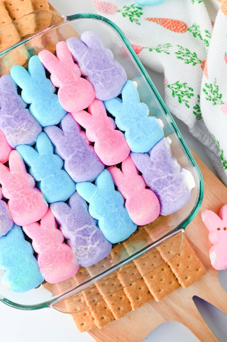 A glass dish containing rows of pink, blue, and purple marshmallow bunnies on top of graham crackers, with a carrot-patterned cloth nearby.