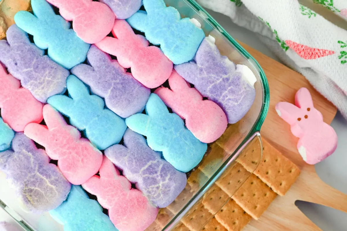 A glass dish filled with pink, blue, and purple marshmallow bunnies arranged in rows. A small pink bunny marshmallow is next to the dish on a wooden surface with graham crackers.