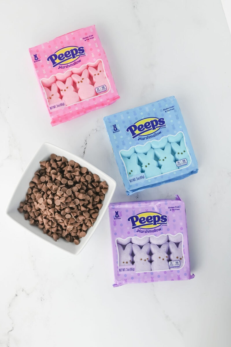 Three packs of Peeps marshmallows in pink, blue, and purple are next to a square bowl filled with chocolate chips on a white surface.