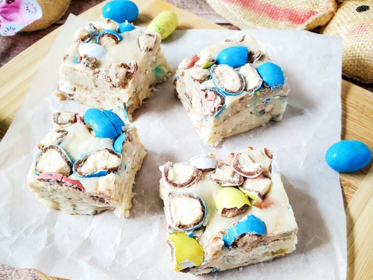 Four square pieces of dessert with colored candies on top, placed on parchment paper on a wooden board, surrounded by decorative items and additional candies.