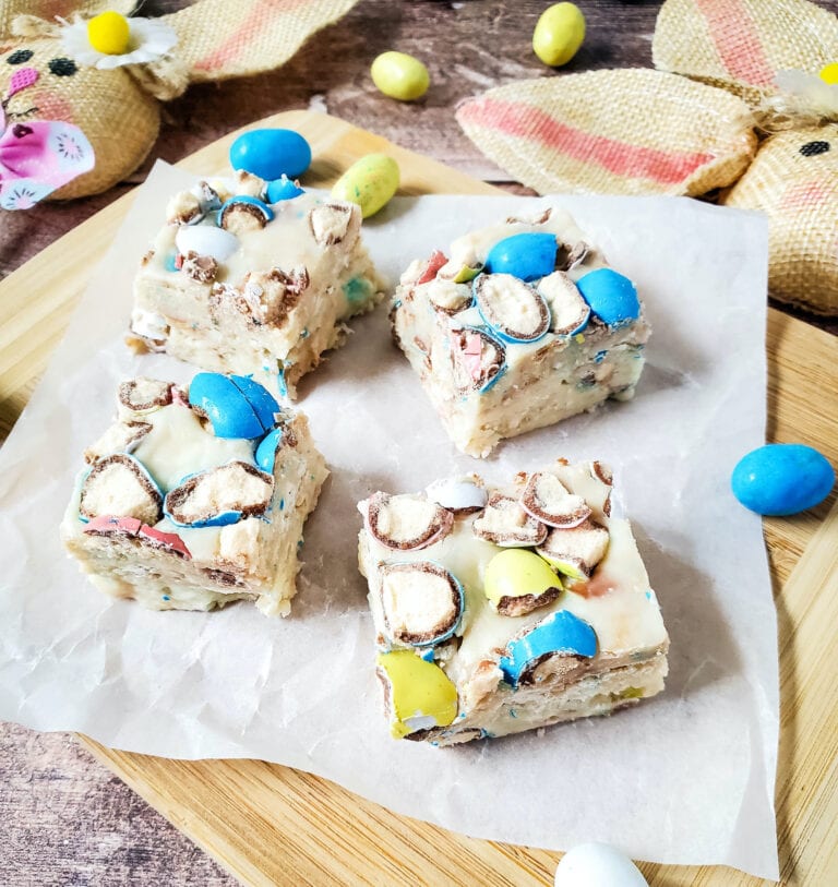 Four square pieces of dessert with colored candies on top, placed on parchment paper on a wooden board, surrounded by decorative items and additional candies.