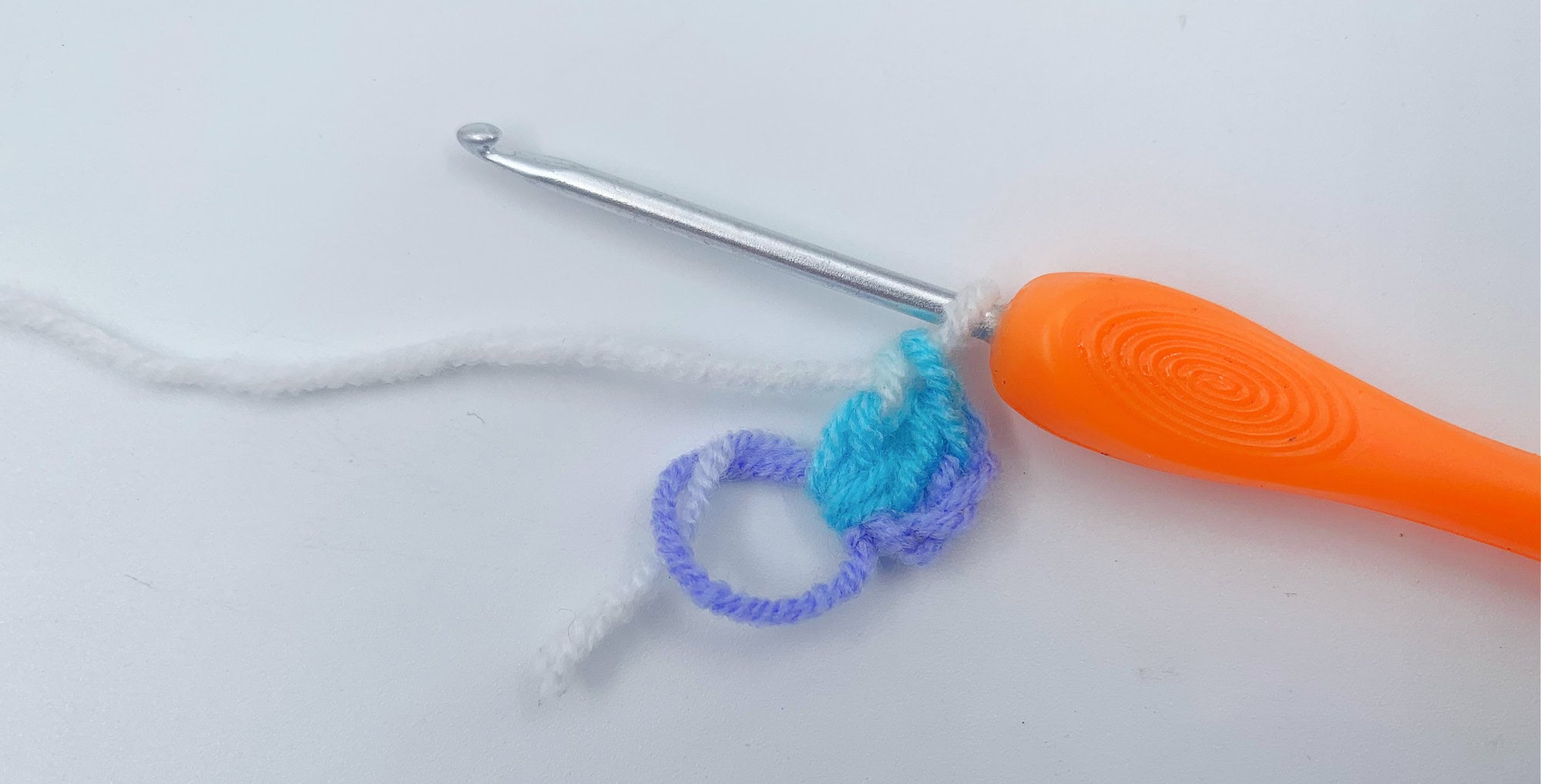 An orange-handled crochet hook with white and purple yarn in progress on a white surface.