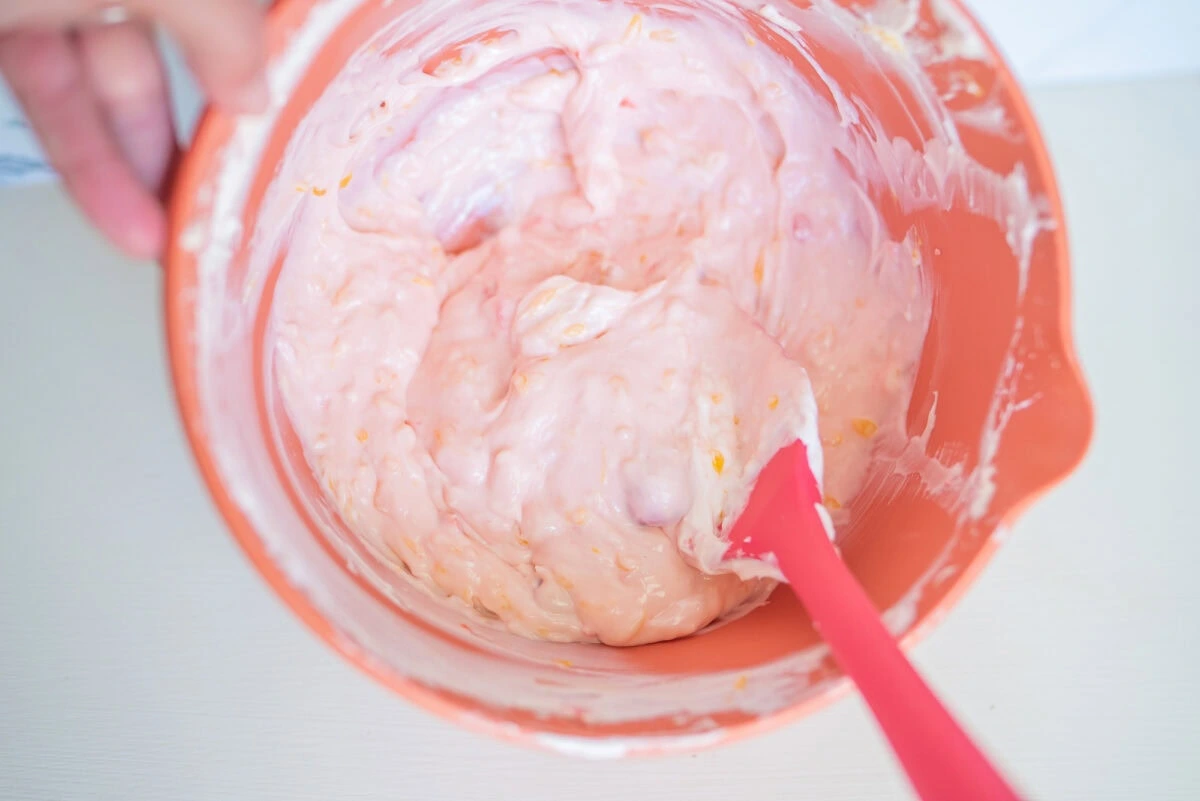 A pink creamy mixture is being stirred in an orange bowl with a red spatula.