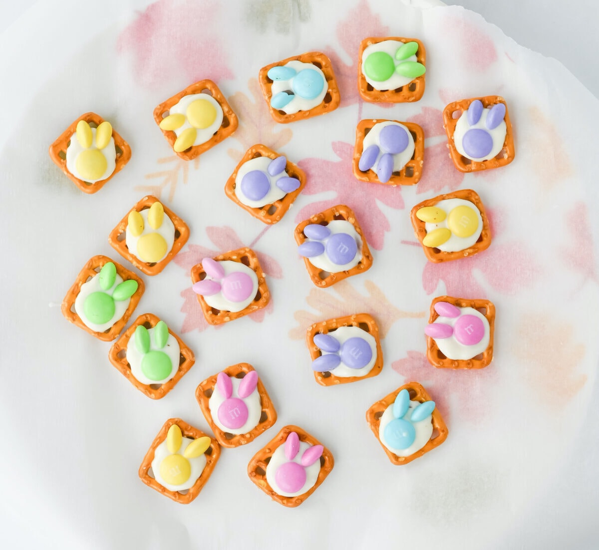 Easter bunny pretzel treats feature square pretzels topped with pastel-colored candies shaped like bunnies, all beautifully arranged on parchment paper with a subtle floral design.