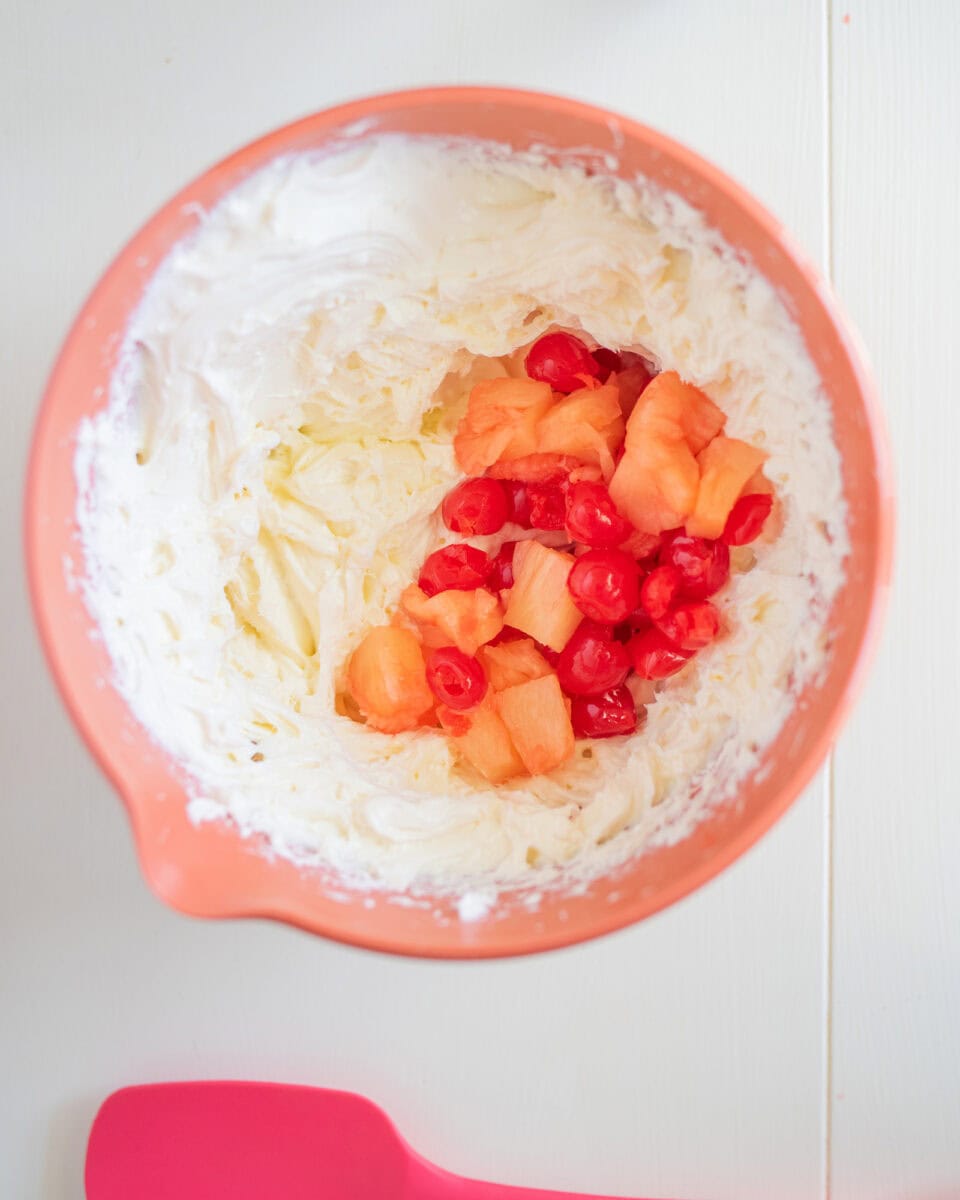 A mixing bowl with whipped cream, diced pineapples, and cherries, beside a pink spatula on a white surface.