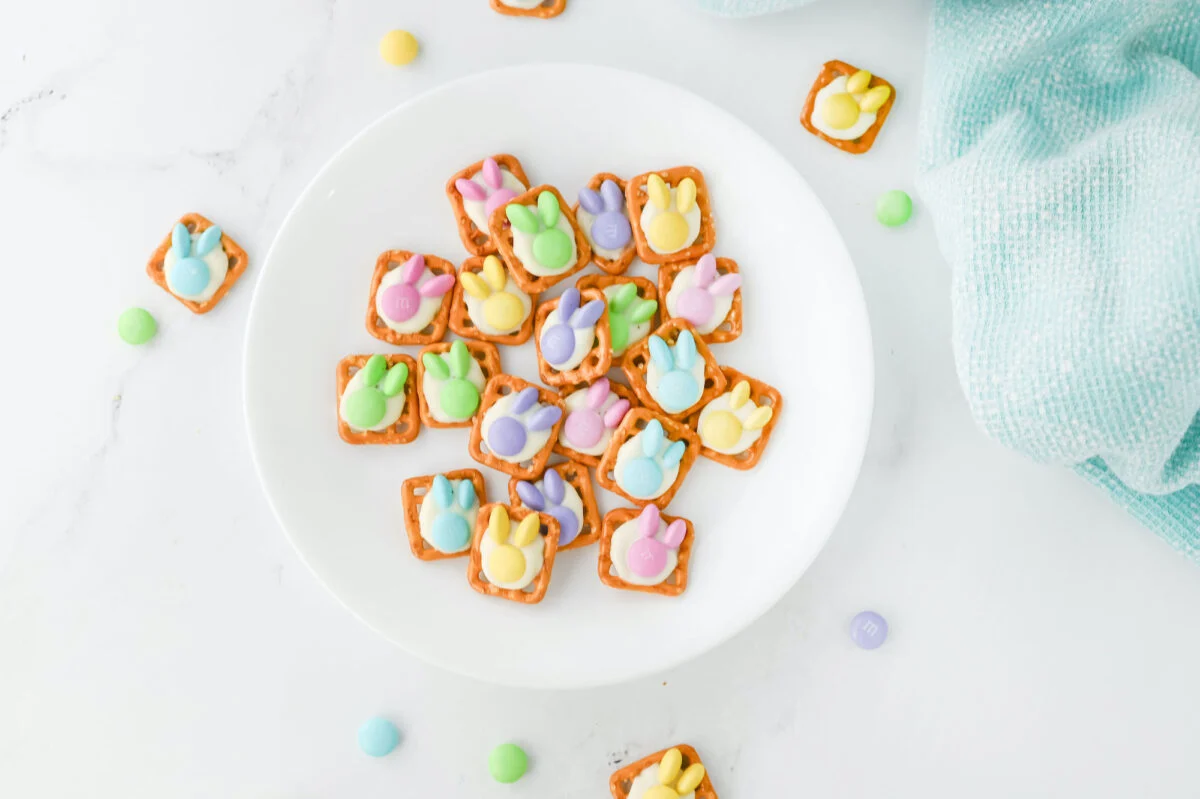 A white plate holds delightful Easter bunny pretzel treats topped with colorful bunny-shaped candies. A light blue cloth and scattered candies add charm to the marble surface.