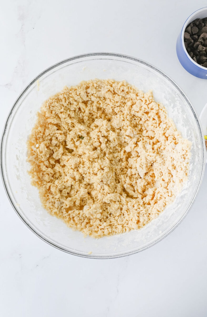 A textured dough mixture in a clear glass bowl on a white surface, with a small bowl of chocolate chips nearby.