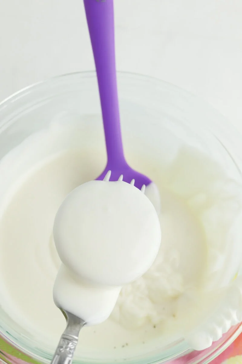 A fork with a purple handle is dipped in a bowl of thick, white liquid, possibly yogurt or cream. A dollop of the liquid is visible on the ends of the fork.