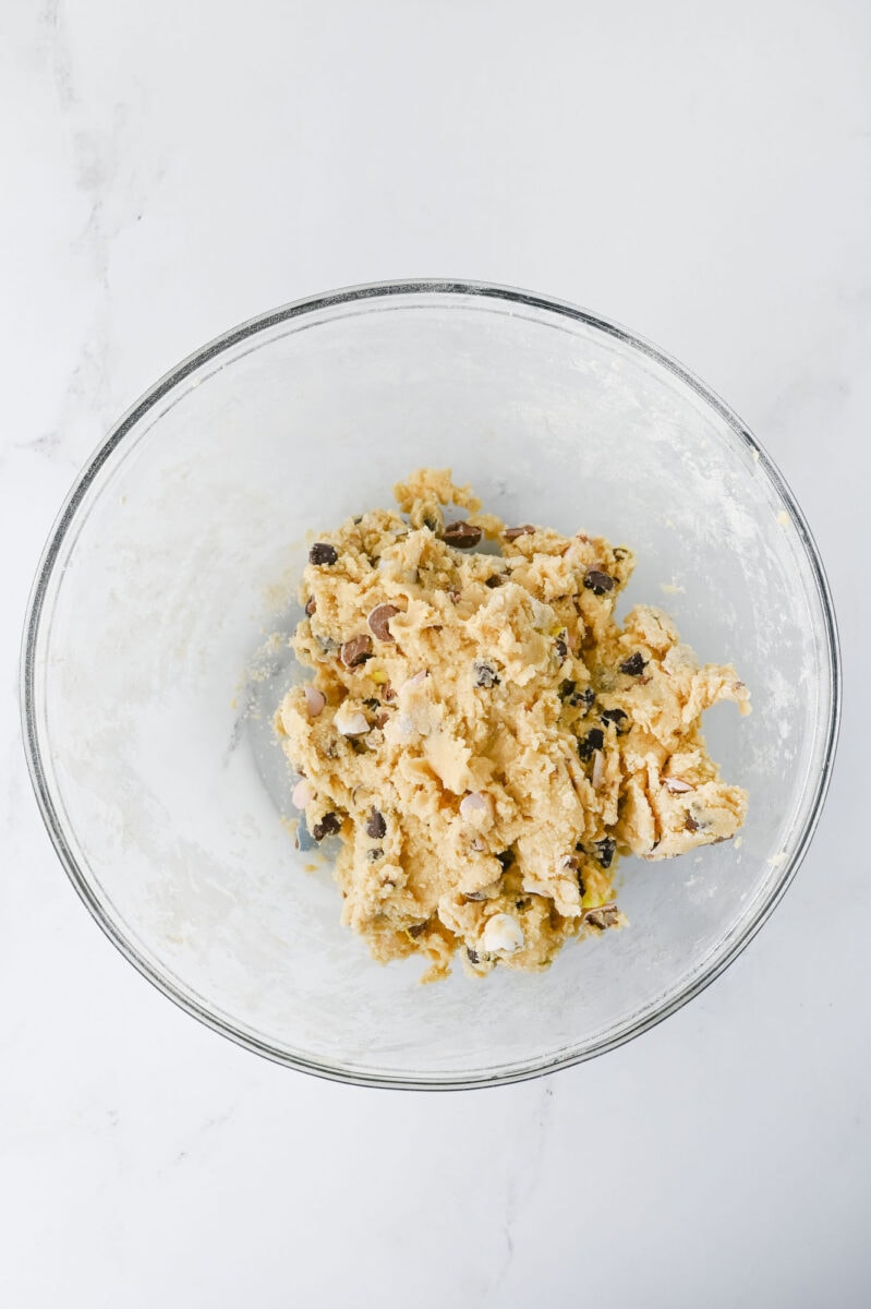 A glass bowl containing raw cookie dough with chocolate chips and chopped nuts on a white marble surface.