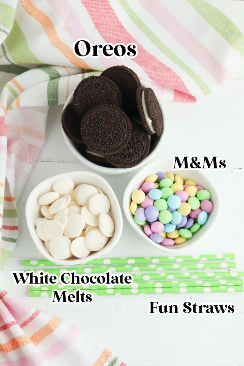 Bowls with Oreos, white chocolate melts, and colorful M&Ms. Green and white polka dot straws are next to the bowls on a white surface with striped cloths.