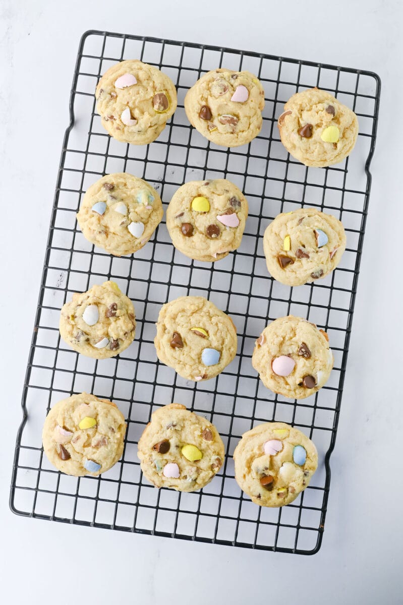 Twelve cookies with pastel-colored candies are arranged on a black cooling rack.