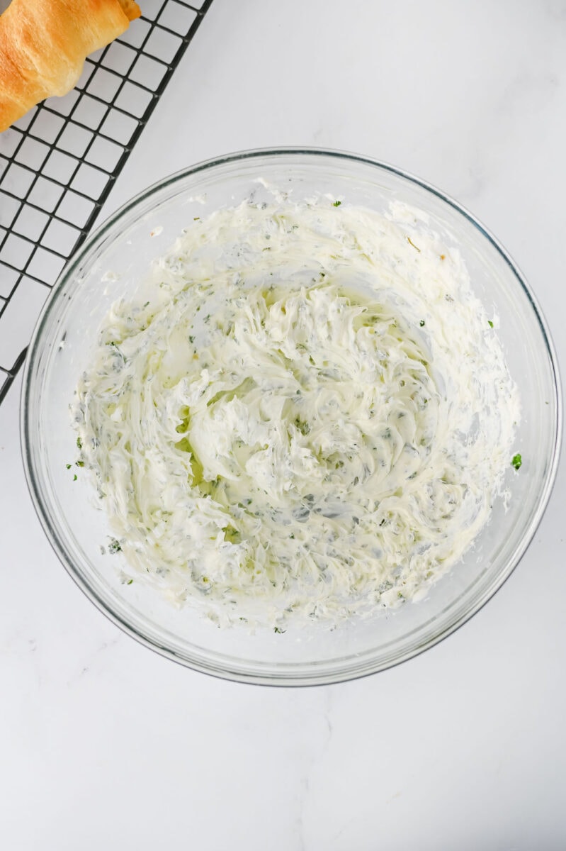 A clear glass bowl filled with creamy herb-infused butter sits on a white countertop. A section of a cooling rack with a croissant is partially visible.