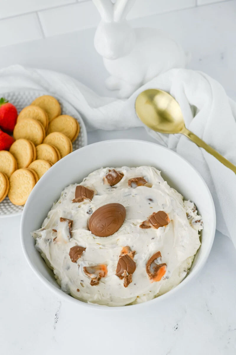 A bowl of white cream cheese dip with chocolate pieces, a central chocolate egg, a plate of cookies, and strawberries nearby. A gold spoon and a white rabbit figure are visible in the background.