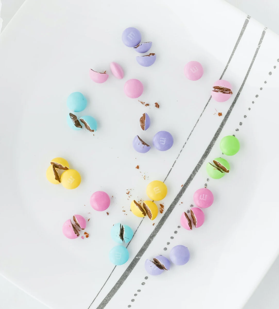 Colorful candy-coated chocolates, reminiscent of Easter bunny pretzel treats, are scattered on a white plate with black lines and dots, some artfully split open.