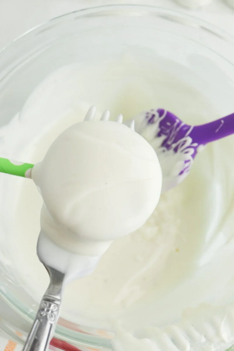 A fork holding a round, white chocolate-covered item over a glass bowl. Nearby is a purple spatula with white patterns.