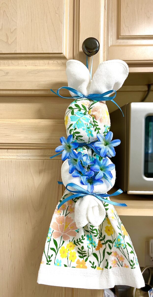 A decorative dish towel bunny, adorned with blue flowers, ribbons, and a floral pattern, hangs charmingly from a cabinet knob in the kitchen.