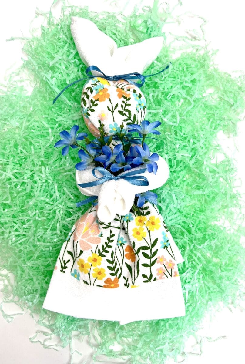 A floral-patterned dish towel folded into a charming bunny, adorned with blue flowers and ribbons, rests gracefully on a bed of green shredded paper.