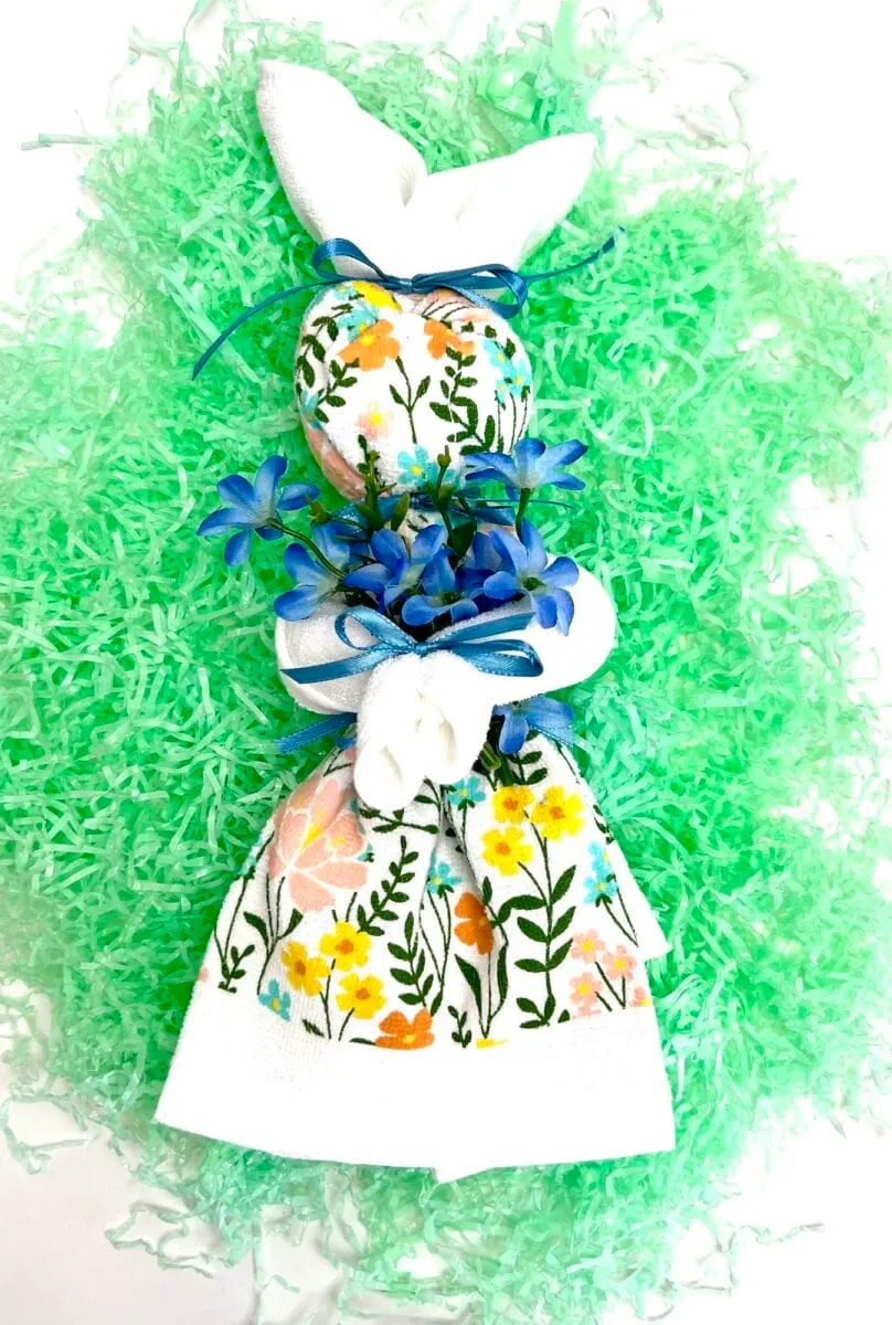 A floral-patterned dish towel folded into a charming bunny, adorned with blue flowers and ribbons, rests gracefully on a bed of green shredded paper.