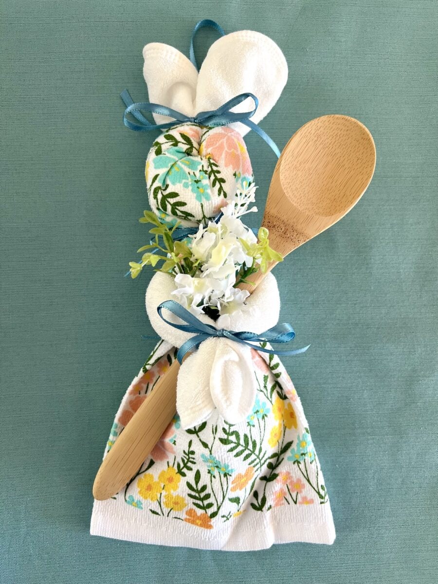 A gift bundle featuring floral-patterned towels with a charming dish towel bunny, a wooden spoon, white flowers, and blue ribbon on a teal background.