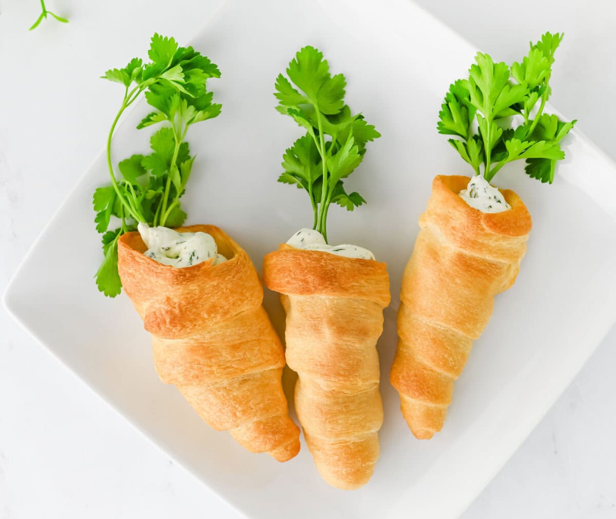 Three crescent roll carrots filled with herb cream cheese, garnished with parsley, sit elegantly on a white square plate.