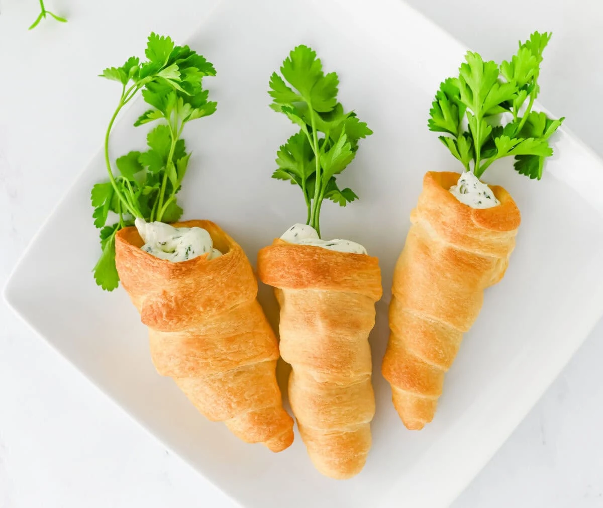 Three crescent roll carrots filled with herb cream cheese, garnished with parsley, sit elegantly on a white square plate.