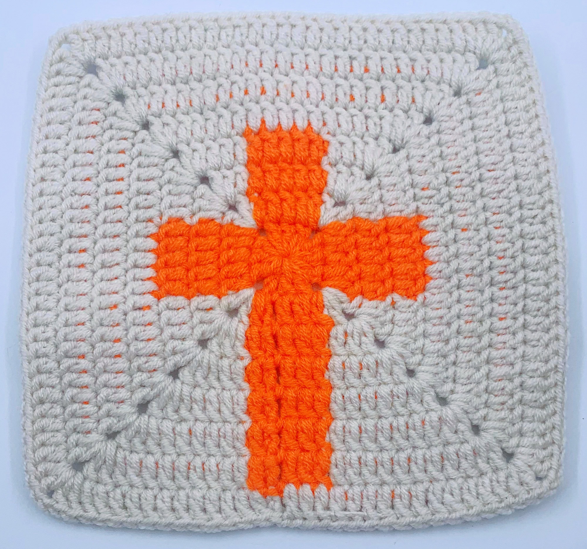 tan crocheted cross granny square with a vibrant orange cross at its center displayed against white backdrop