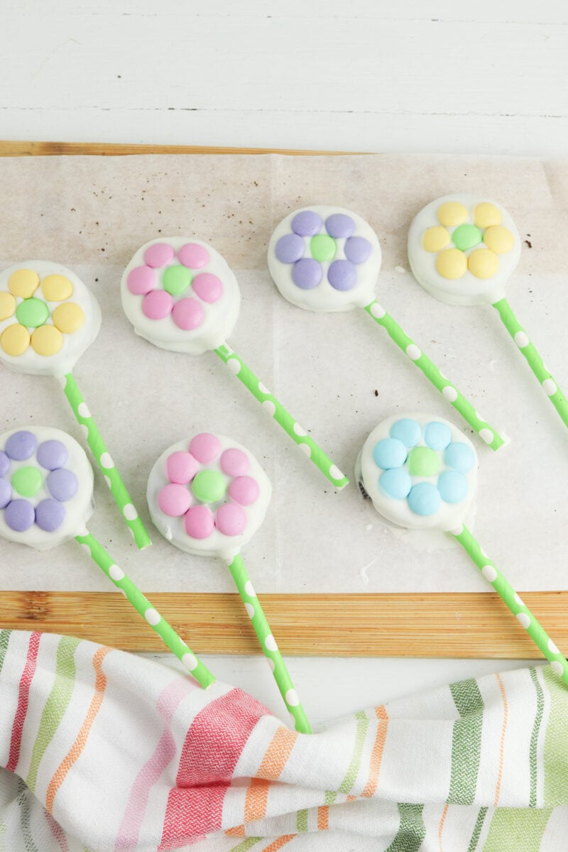 Flower-shaped cake pops with pastel-colored petals on green polka-dotted sticks, placed on parchment paper with a striped cloth nearby.