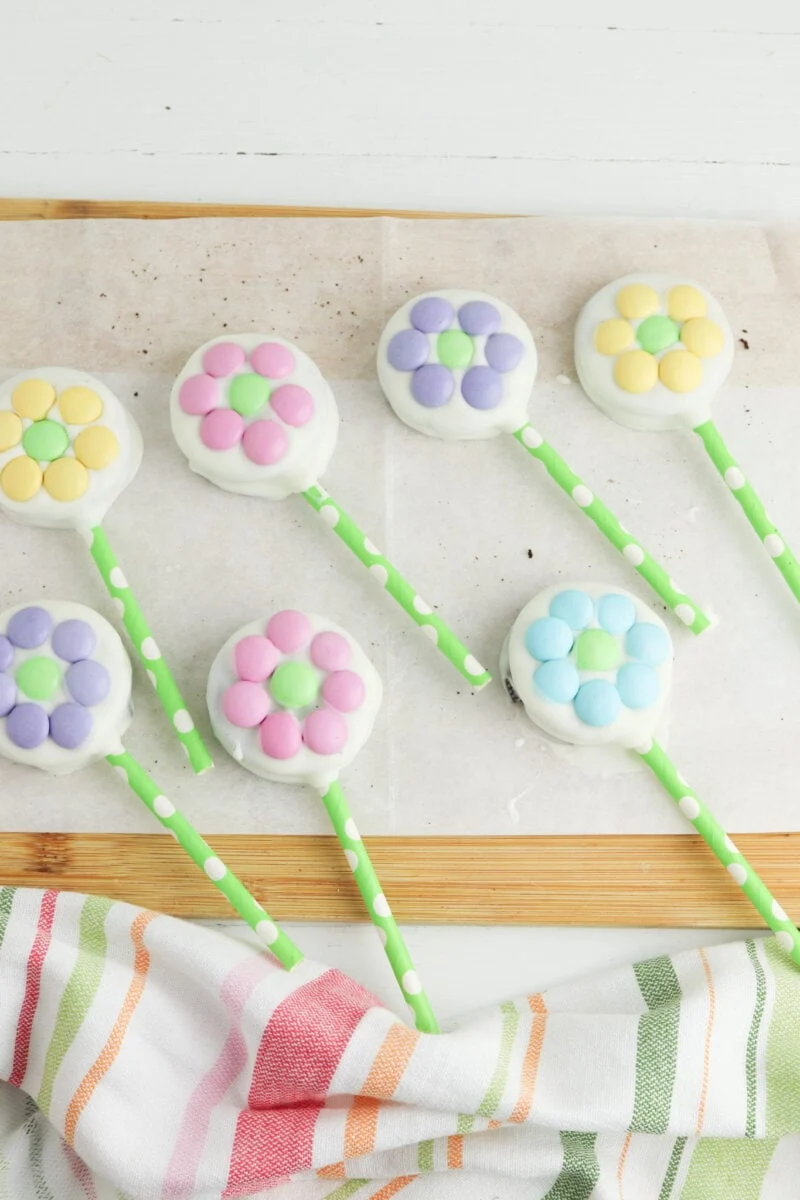 Flower-shaped cake pops with pastel-colored petals on green polka-dotted sticks, placed on parchment paper with a striped cloth nearby.