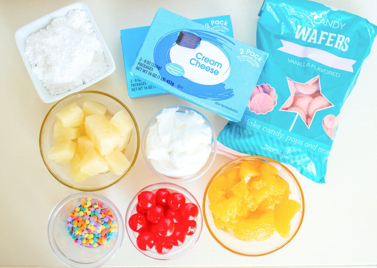 Ingredients for a sweet Easter dip: powdered sugar, cream cheese, vanilla wafers, crushed pineapple, whipped topping, maraschino cherries, mandarin oranges, and colorful sprinkles in bowls.