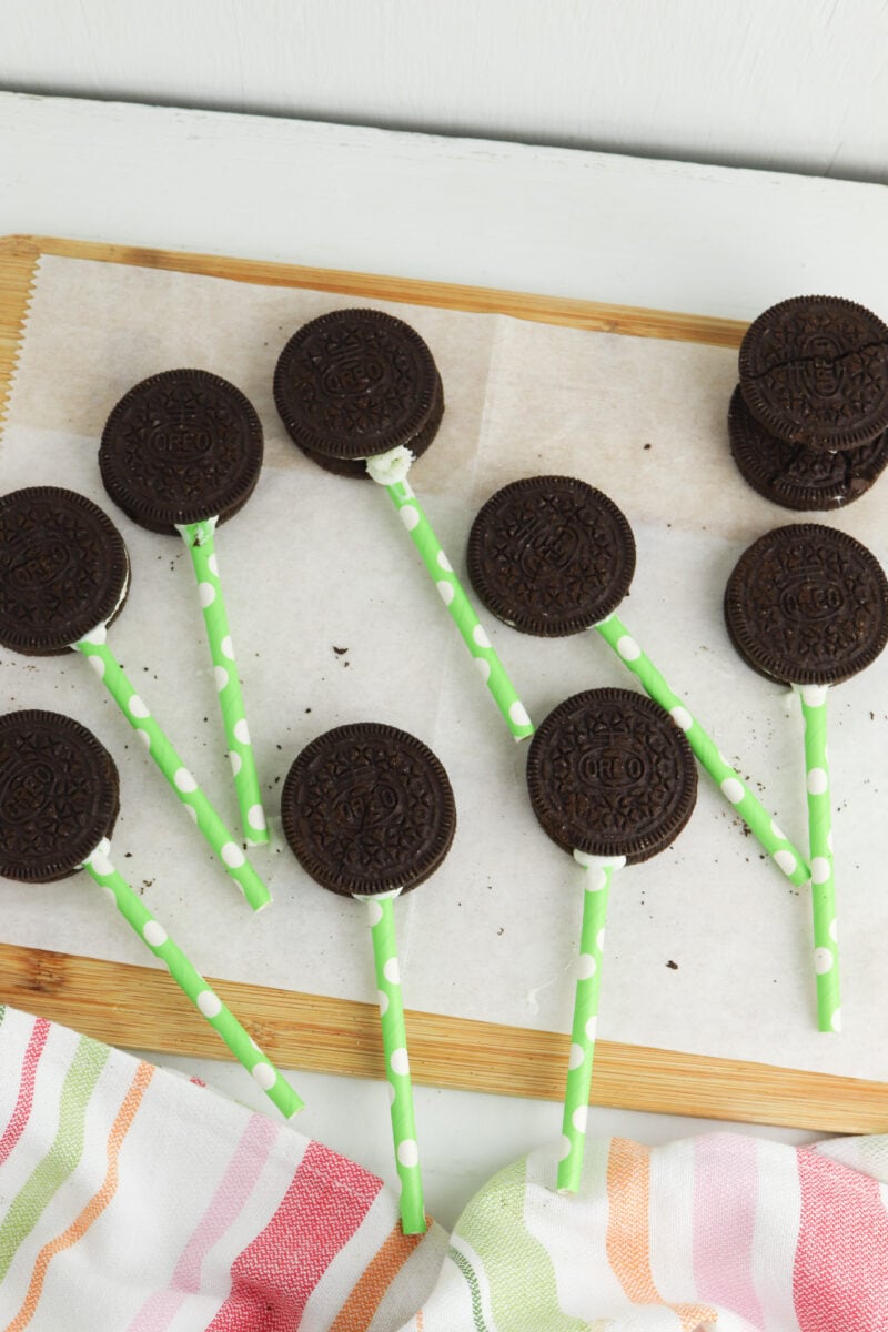 Chocolate sandwich cookies on green polka dot sticks arranged on parchment paper, with a striped cloth in the corner.