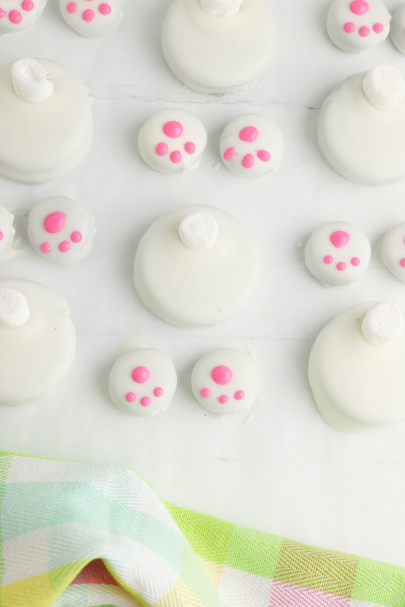 White cookies with pink paw prints and a small white dot on top, arranged on a light surface. A pastel plaid cloth is partially visible at the bottom.