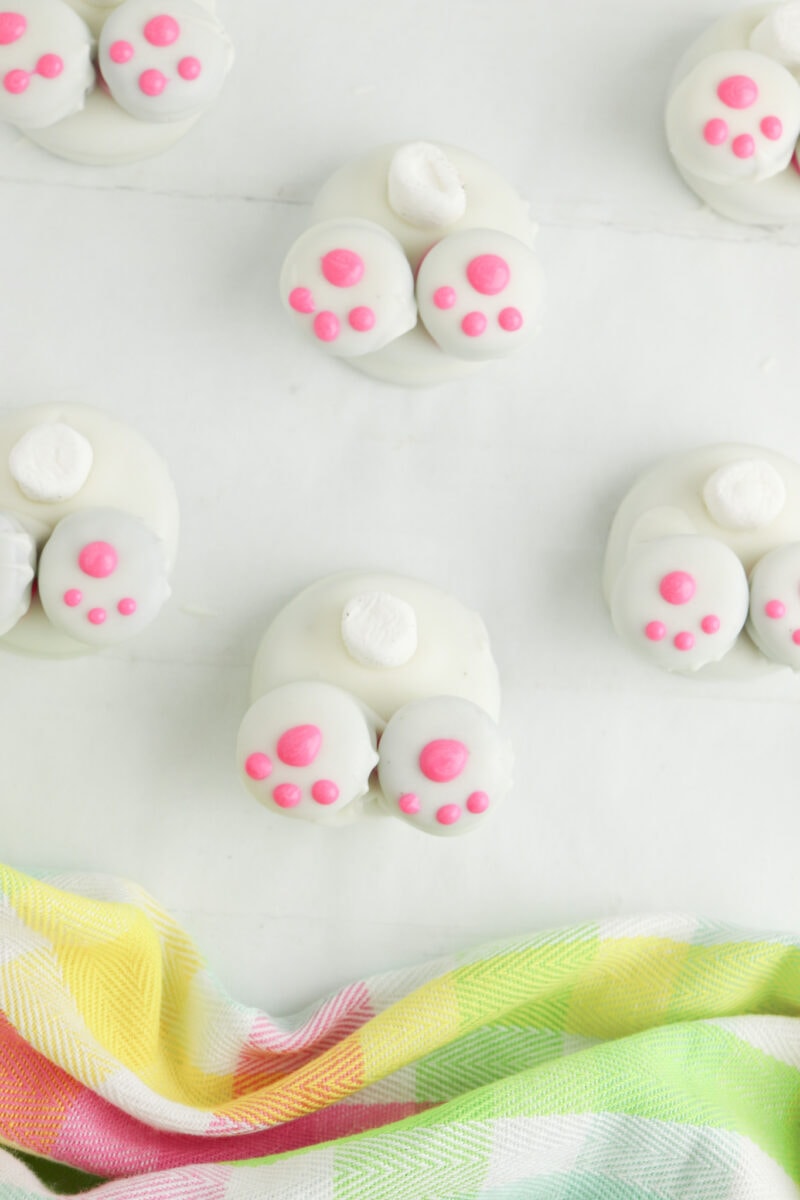 White chocolate treats decorated to resemble bunny feet, with pink paw prints and white tails, arranged on a light surface next to a colorful, patterned cloth.