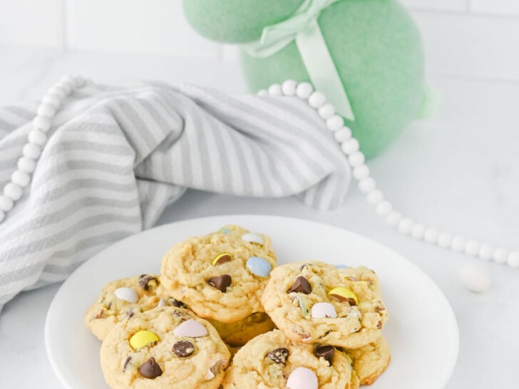 A plate of cookies with chocolate chips and Cadbury eggs sits on a white surface. A gray striped cloth and a green felt rabbit decoration are in the background.