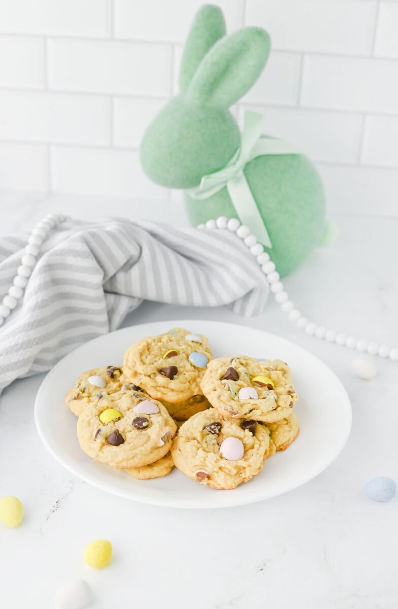 A plate of cookies with chocolate chips and pastel candies sits on a white surface. A gray striped cloth and a green felt rabbit decoration are in the background.