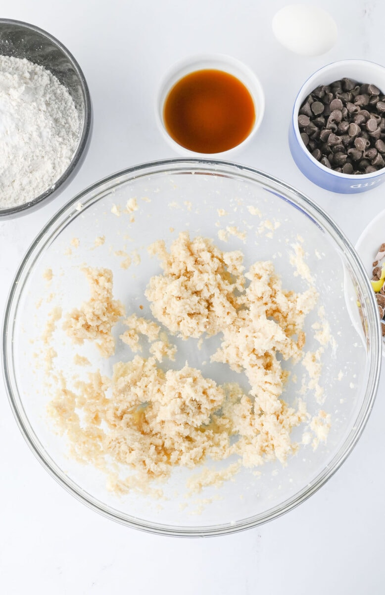 Glass bowl with cookie dough mixture, surrounded by ingredients: flour, vanilla extract, chocolate chips, and an egg on a white surface.