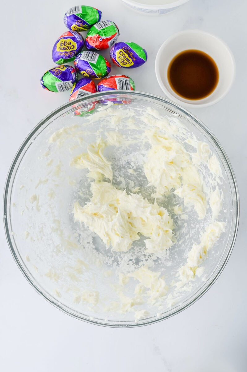 A glass bowl with creamy, whipped mixture, a small bowl of brown liquid, and several Cadbury Creme Eggs on a white surface.