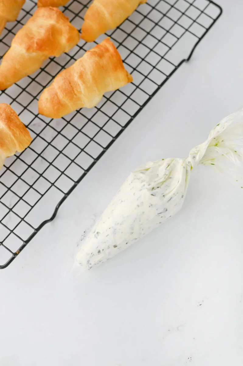 Golden pastry shells on a cooling rack next to a piping bag filled with herbed cream mixture on a white surface.