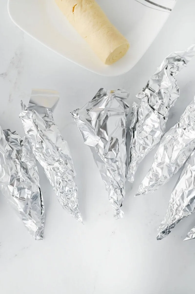 Several foil-wrapped cones are placed on a light surface, with a roll of dough on a white dish in the background.