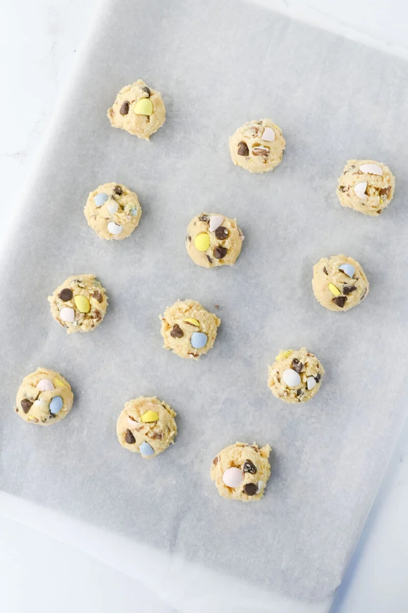 Cookie dough balls with chocolate chips and colorful candies on a parchment-lined baking sheet.