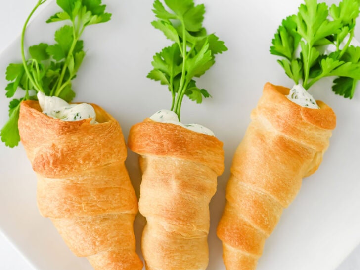 Three crescent roll carrot-shaped pastries filled with creamy topping and garnished with fresh parsley on a white plate.