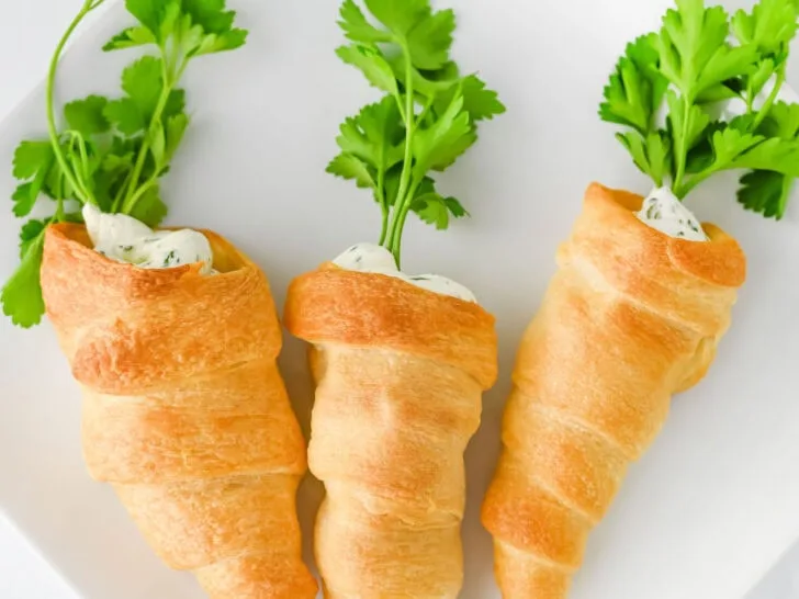 Three crescent roll carrot-shaped pastries filled with creamy topping and garnished with fresh parsley on a white plate.