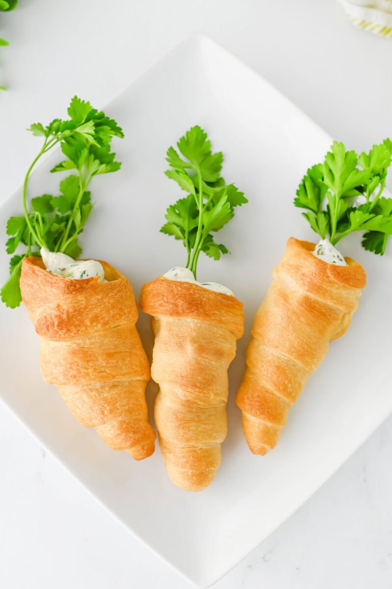 Three crescent roll carrot-shaped pastries filled with creamy topping and garnished with fresh parsley on a white plate.