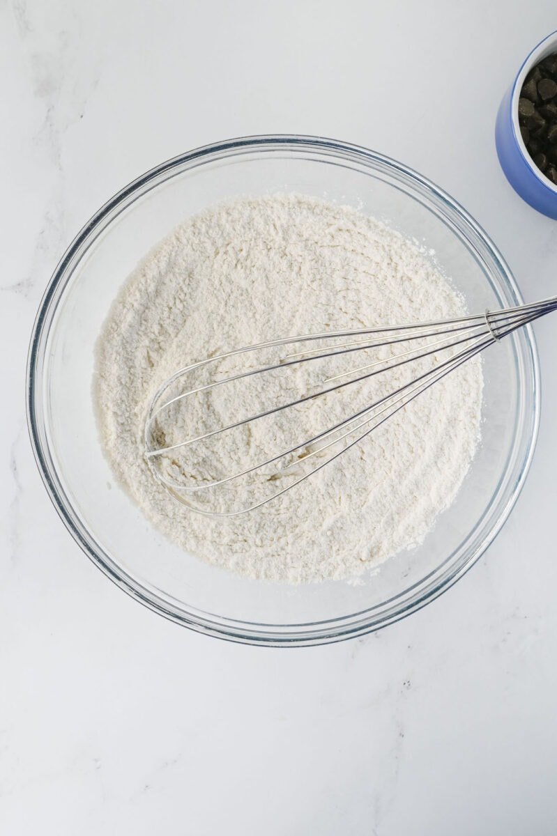 A glass bowl contains a white flour mixture with a metal whisk inside. A small blue bowl with dark contents is partially visible in the corner.