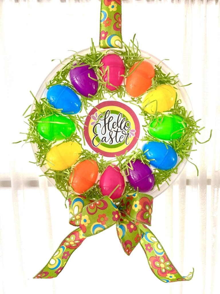 A colorful Easter wreath made of plastic eggs and green grass with a "Hello Easter" sign in the center. A decorative ribbon with a floral pattern is attached at the bottom.