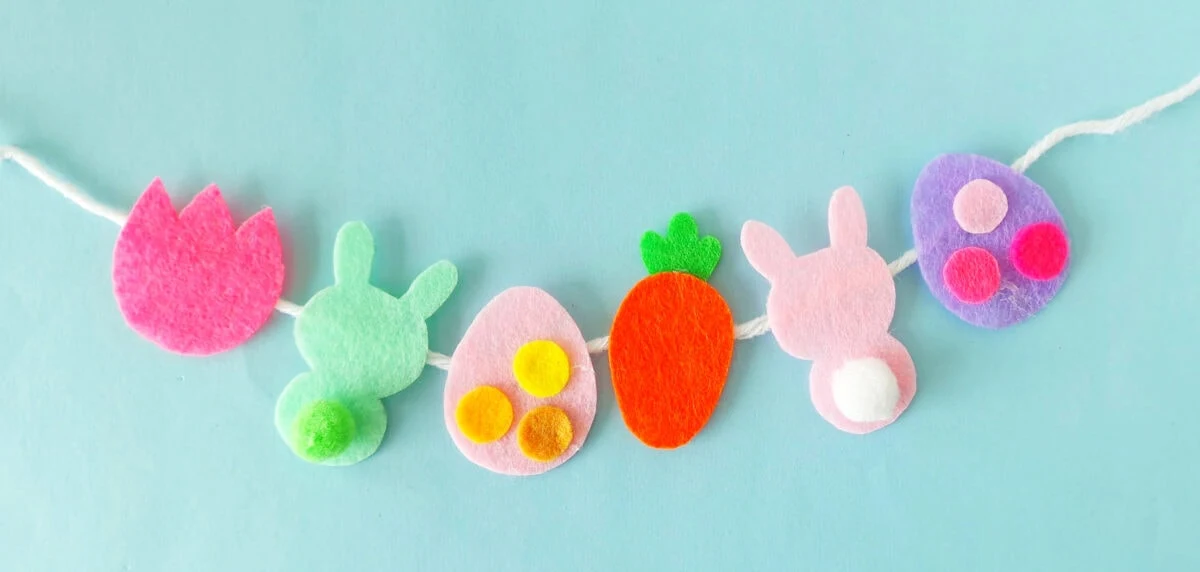 A festive garland of felt shapes on a string against a light blue background, featuring a pink flower, two bunny silhouettes, two eggs, and an orange carrot.