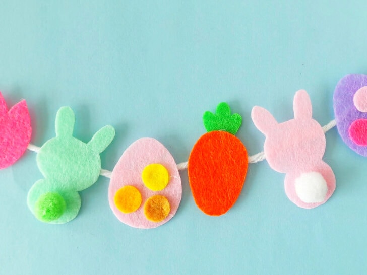 A festive garland of felt shapes on a string against a light blue background, featuring a pink flower, two bunny silhouettes, two eggs, and an orange carrot.