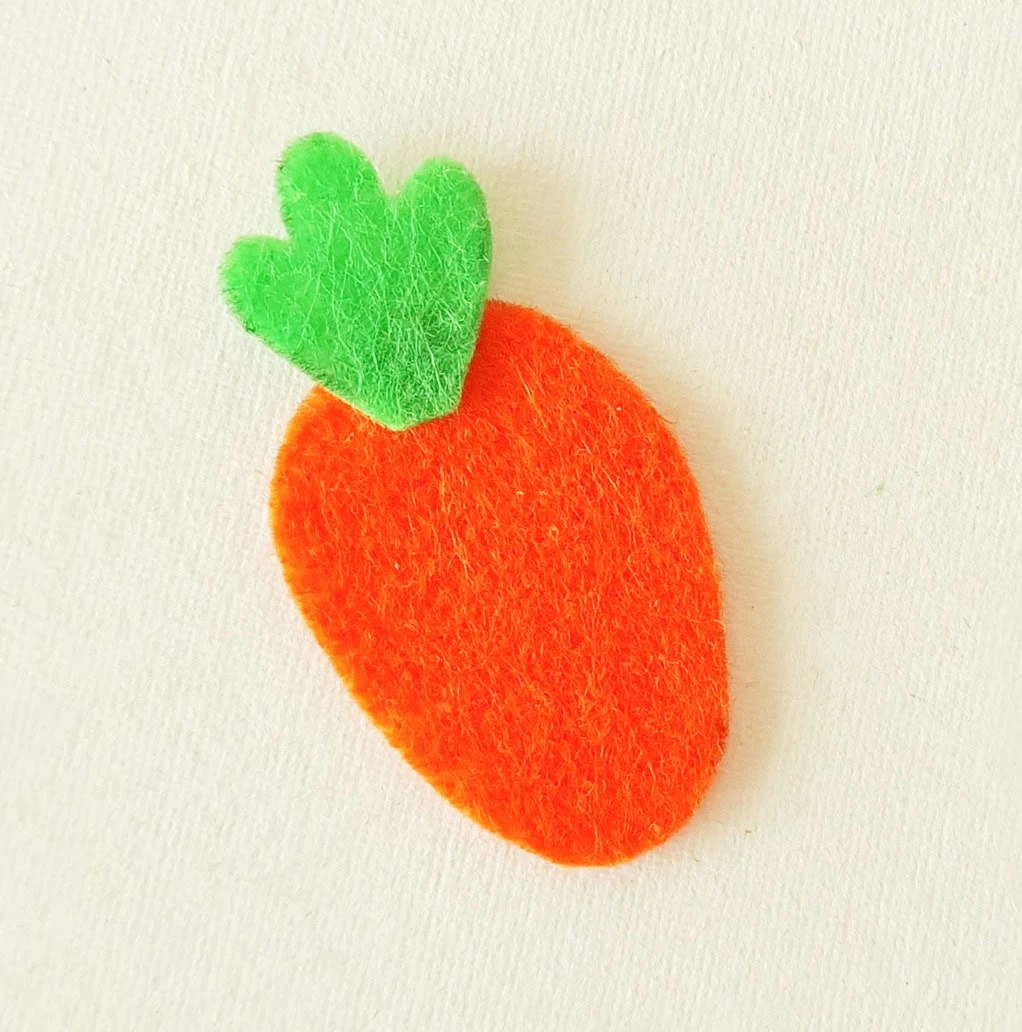 Felt cutout of an orange carrot with green leaves on a white background.