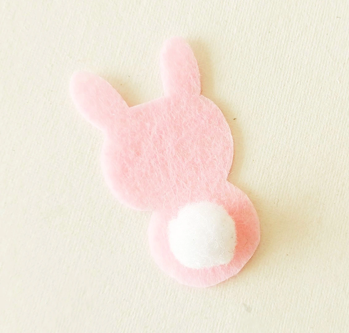 A pink felt cutout of a bunny shape with a white pom-pom attached to its base, representing a tail.