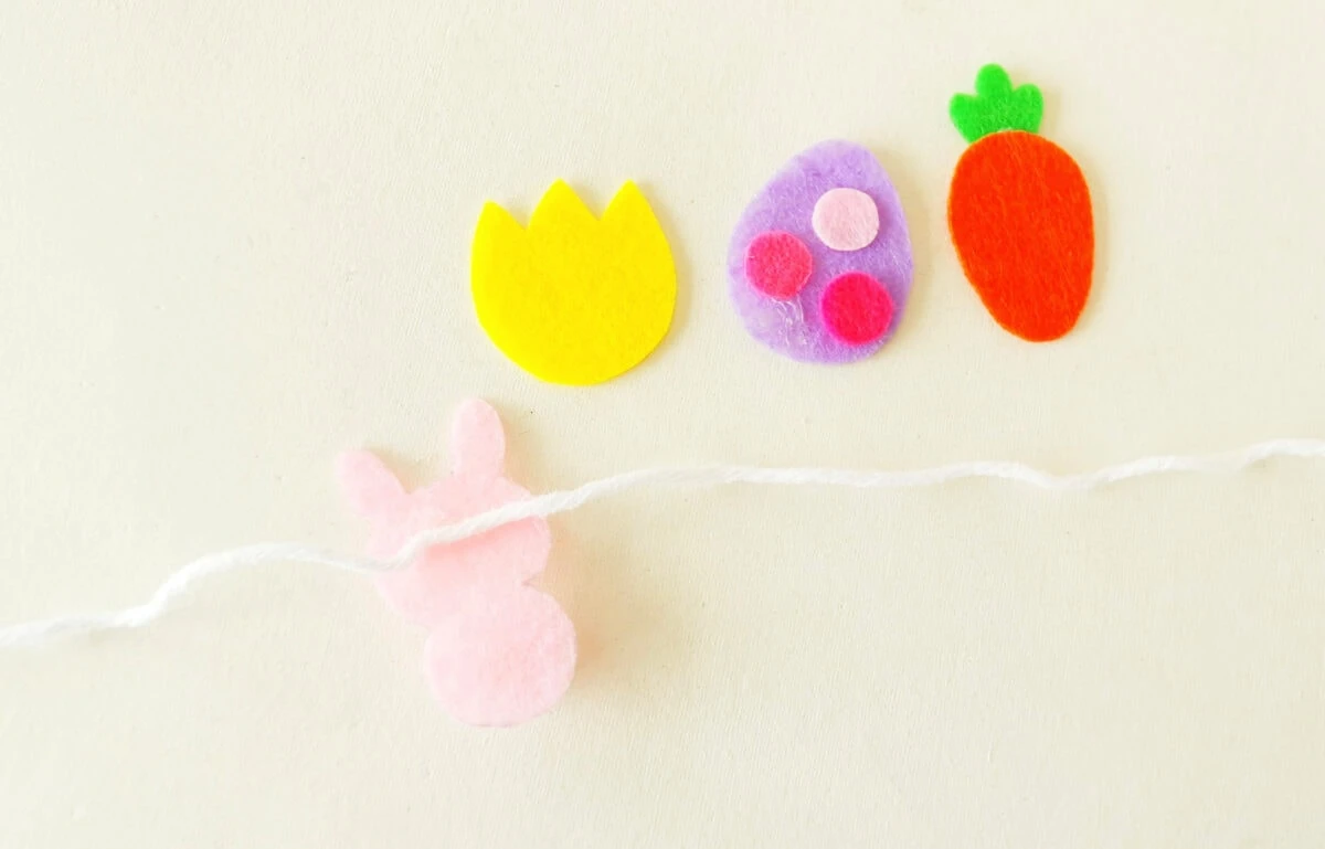 Colorful felt shapes of a yellow tulip, purple egg, orange carrot, and pink bunny on string against a light background.