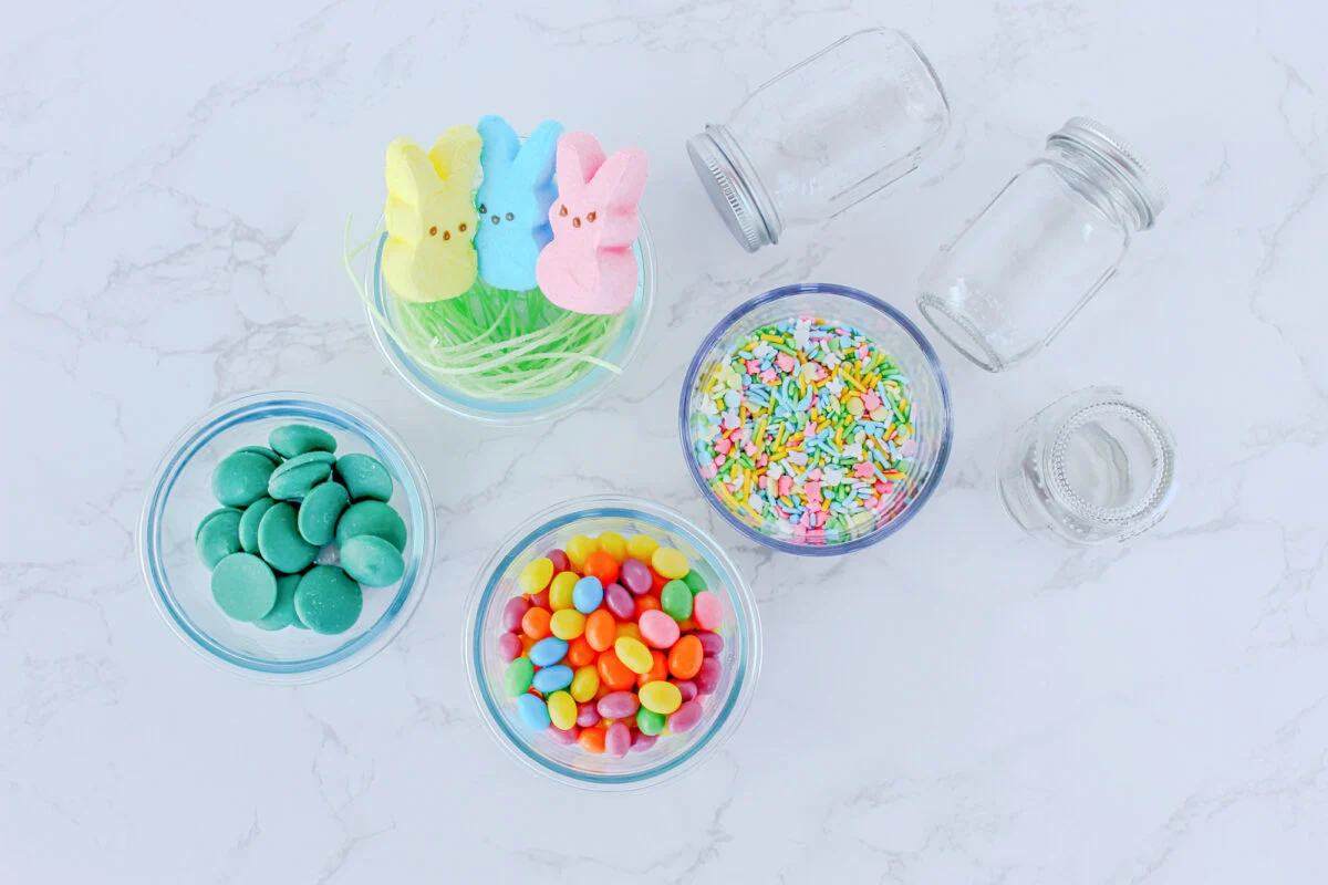 Five glass containers hold various candies: chocolate melts, colorful jelly beans, pastel marshmallow bunnies, rainbow sprinkles, and two empty jars on a marble surface.